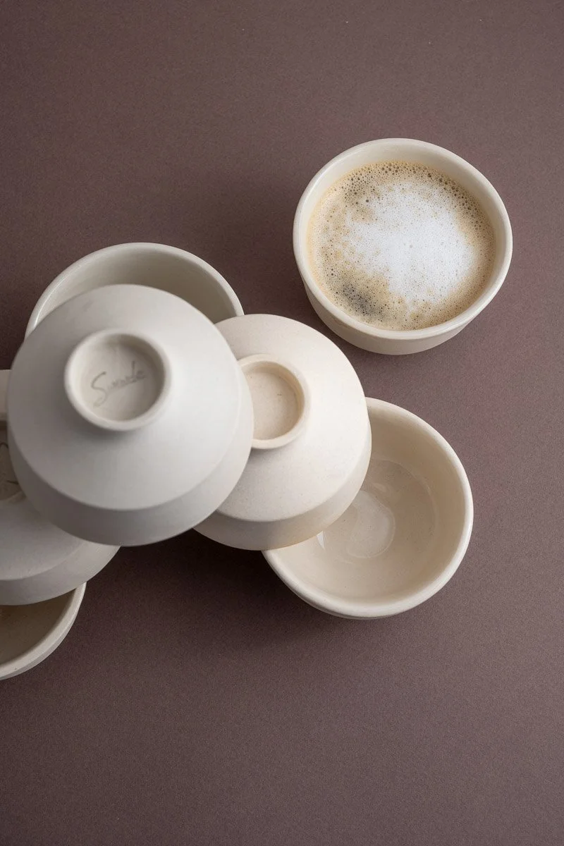 A top-down view of several white ceramic cups and bowls on a purple surface, with one cup filled with frothy coffee.