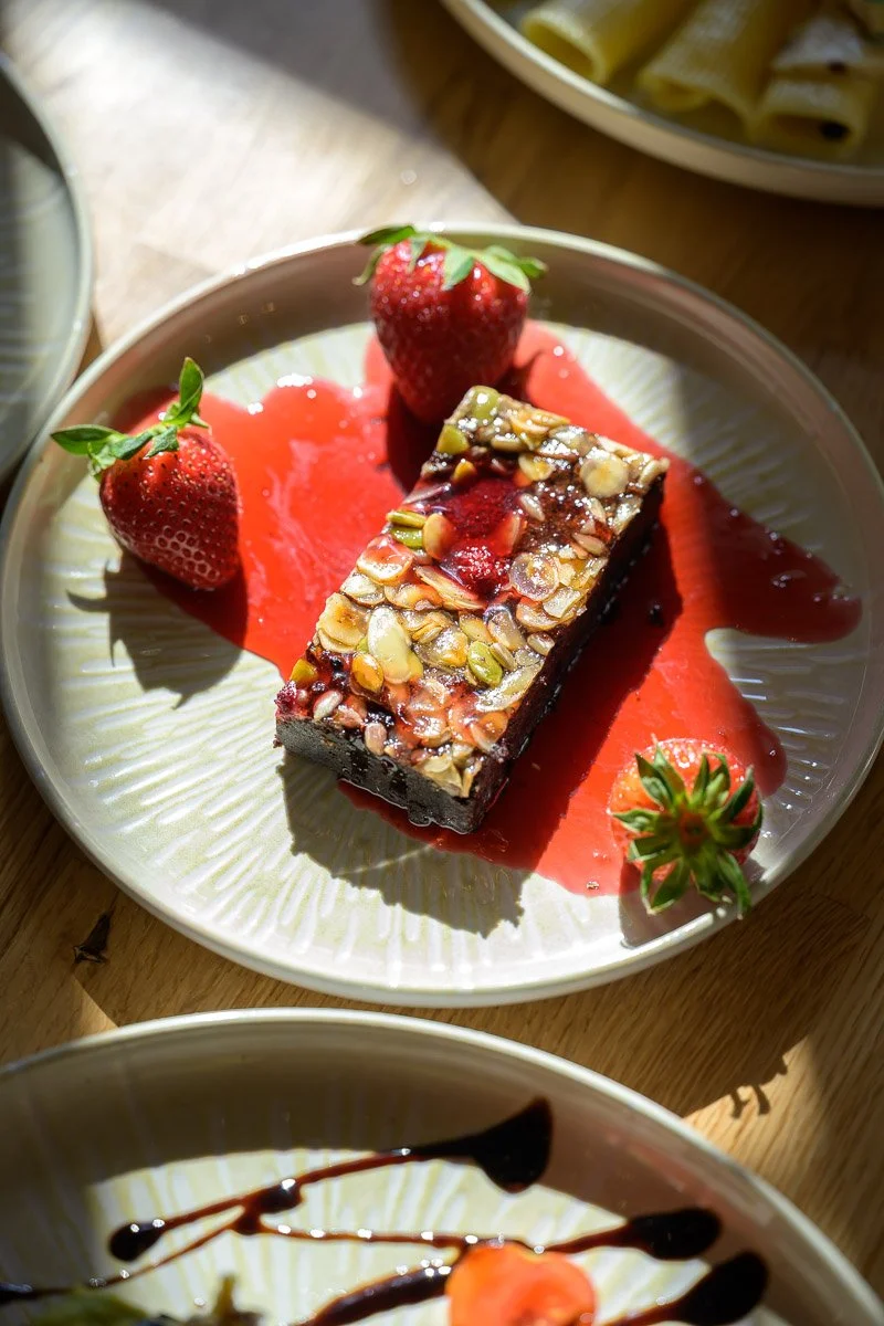 A dessert plate with a brownie topped with nuts, fresh strawberries, and strawberry sauce, served on a white plate with a ridged pattern.