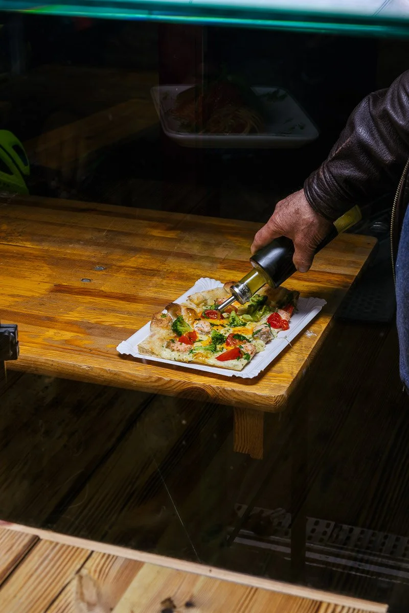 Person adding hot sauce to a slice of pizza on a wooden table.