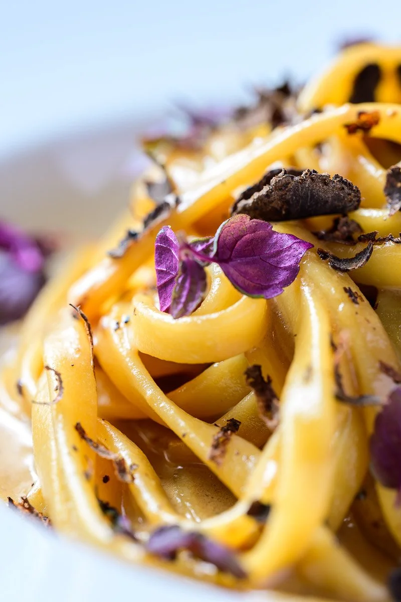 Close-up of yellow pasta topped with purple edible flowers and black truffle shavings.