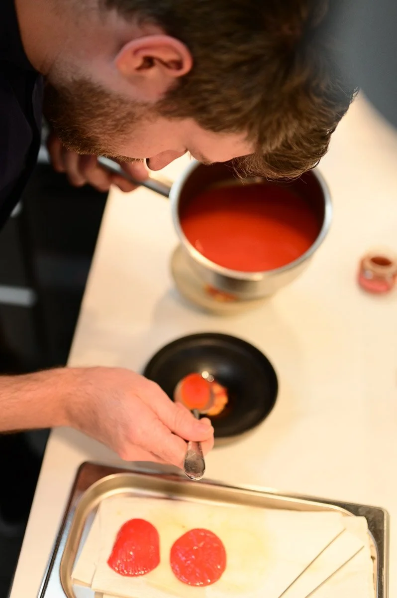 A person with glasses and brown hair is pouring red sauce from a small black dish onto a black plate using a spoon. On the white countertop below, there are two bright red, glossy, round objects, possibly candies or decorative food items, on a piece 