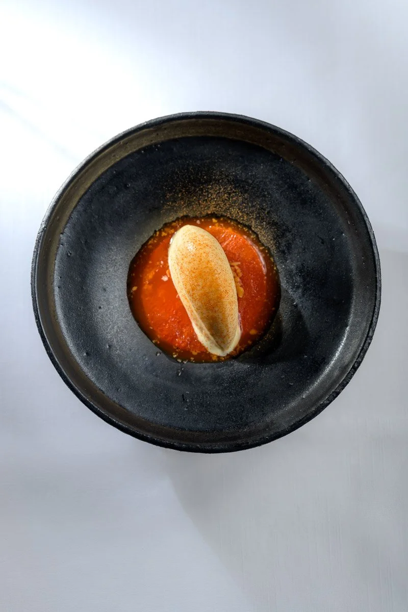 A black bowl containing a red sauce or puree, topped with a dollop of cream and sprinkled with a seasoning or spice.