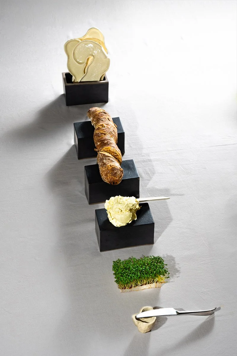 A black rectangular block with cookies, a breadstick, a serving of butter, a sprig of green herbs, and a spoon with a dollop of butter on a gray surface.