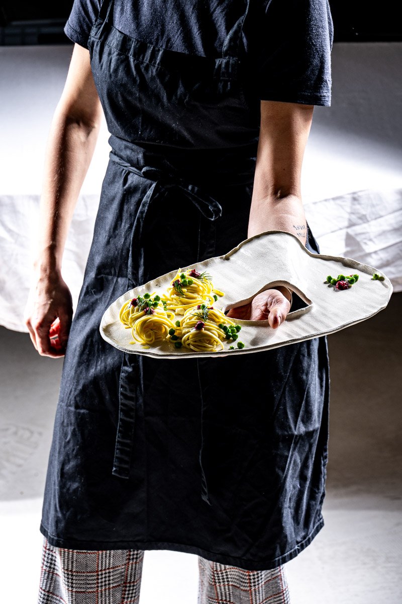 Waiter holding a ceramic platter with three nests of yellow pasta garnished with herbs and small red and green edible flowers, in a restaurant setting.