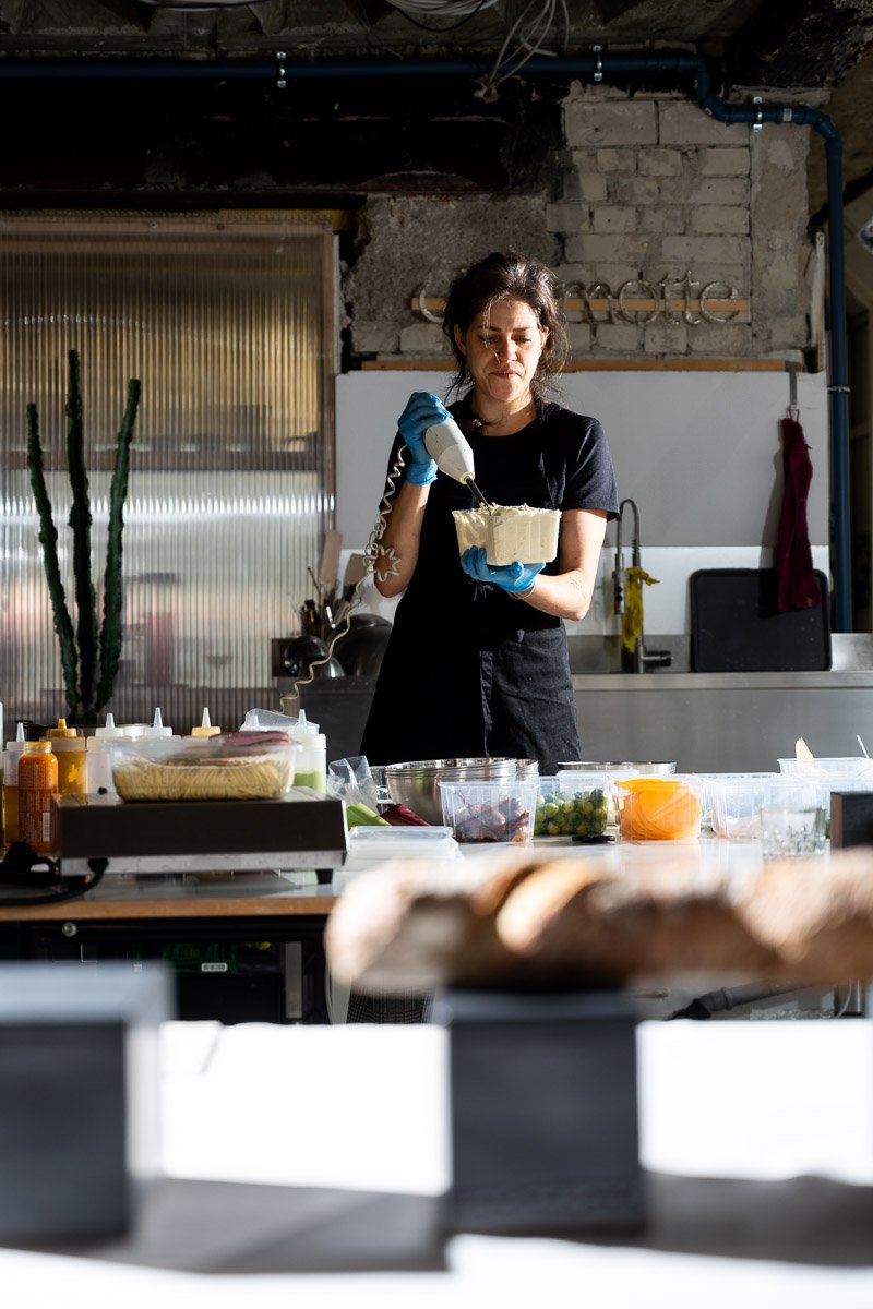 A woman in black apron and blue gloves is using a piping bag to decorate a cake or dessert in a kitchen with various ingredients and tools on the counter.