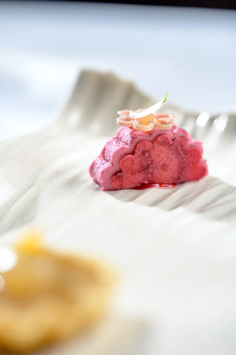 Close-up of a pink ice cream with a small white flower on top, served on a white surface.
