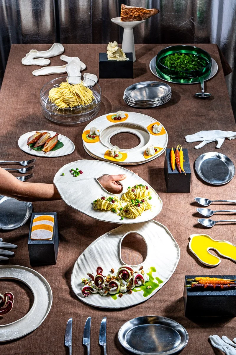 A table set for a culinary display or demonstration with various plates and dishes, including pasta, garnishes, and decorative elements, with a person placing pasta on a white plate and various utensils and serving ware arranged around.