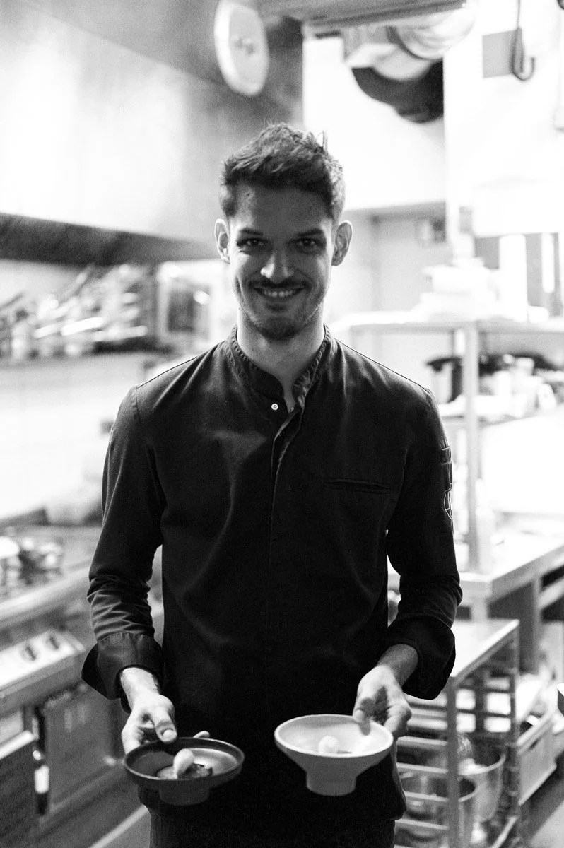 A male chef with a smile holding two bowls in a professional kitchen.