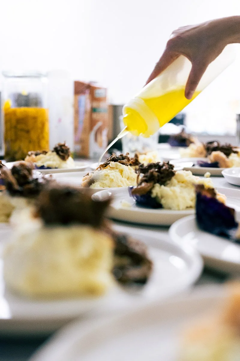 Person squeezing yellow mustard onto mashed potatoes with gravy and cooked meat on plates.