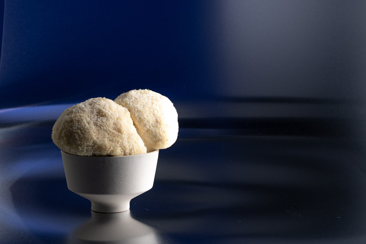 Three scoops of white ice cream in a small white bowl placed on a dark reflective surface with a dark blue background.