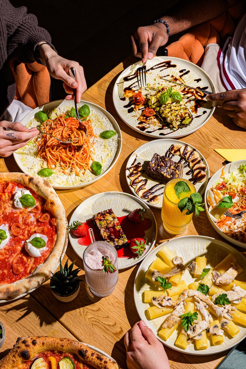A table filled with various dishes including pizza, pasta, salads, desserts, and drinks, with several people eating.