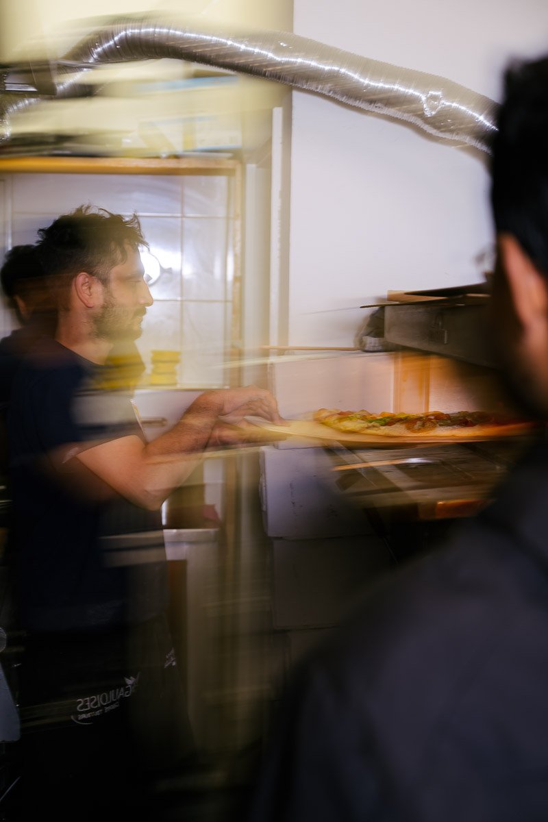 A person with dark hair and a beard making a pizza in a kitchen, seen through a glass or plastic barrier.