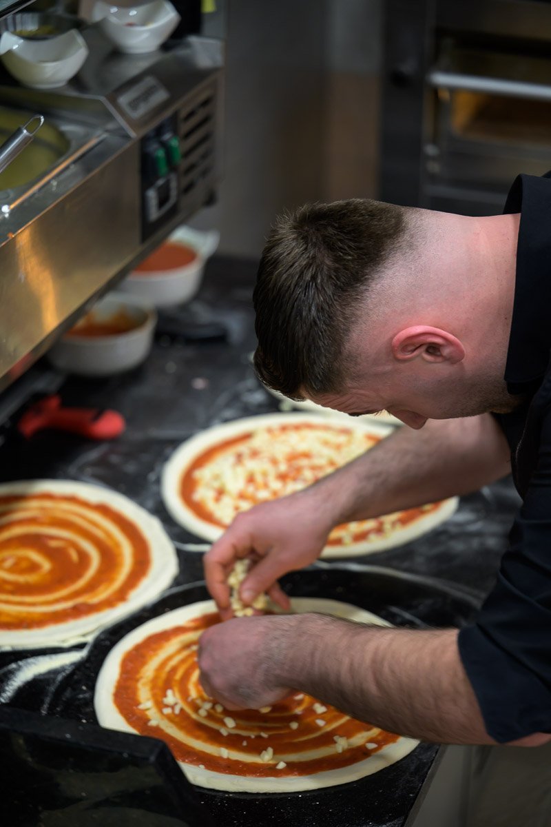 A chef preparing pizzas with tomato sauce and cheese on pizza dough in a commercial kitchen.