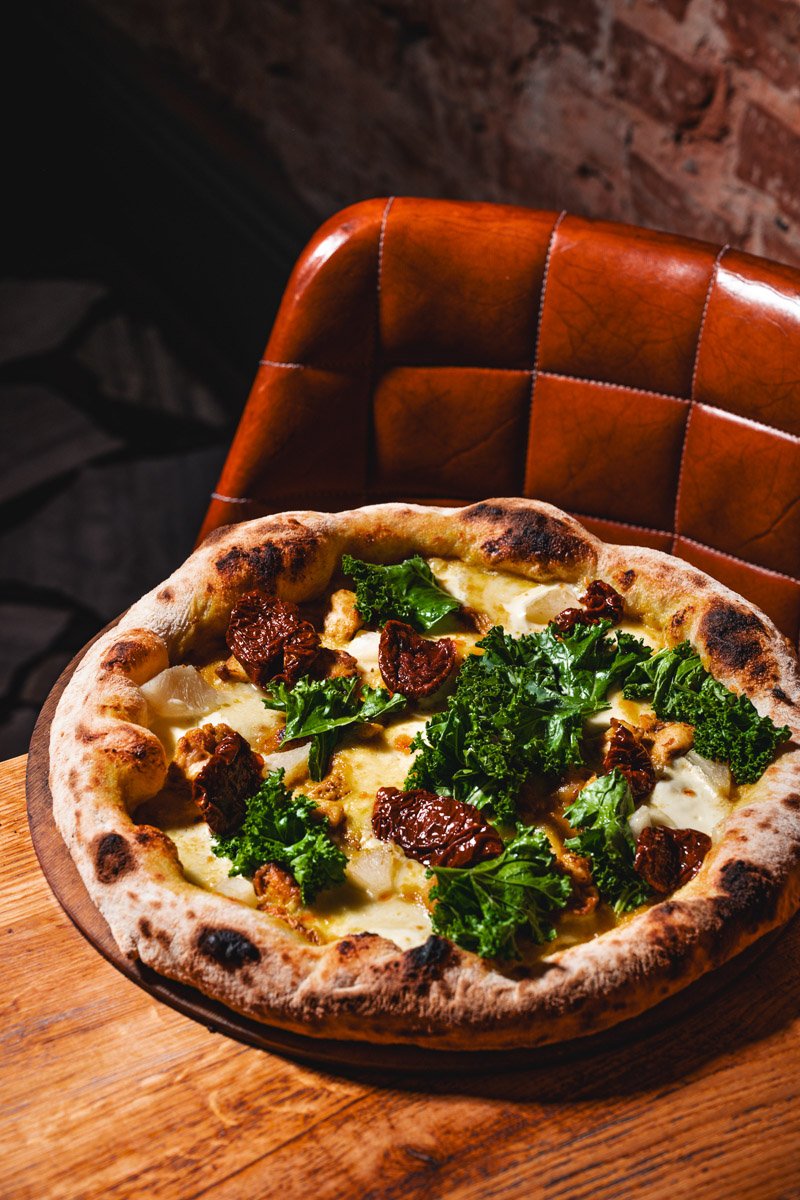 A pizza with cheese, kale, sun-dried tomatoes, and chicken on a wooden table in front of a brown leather chair against a brick wall.