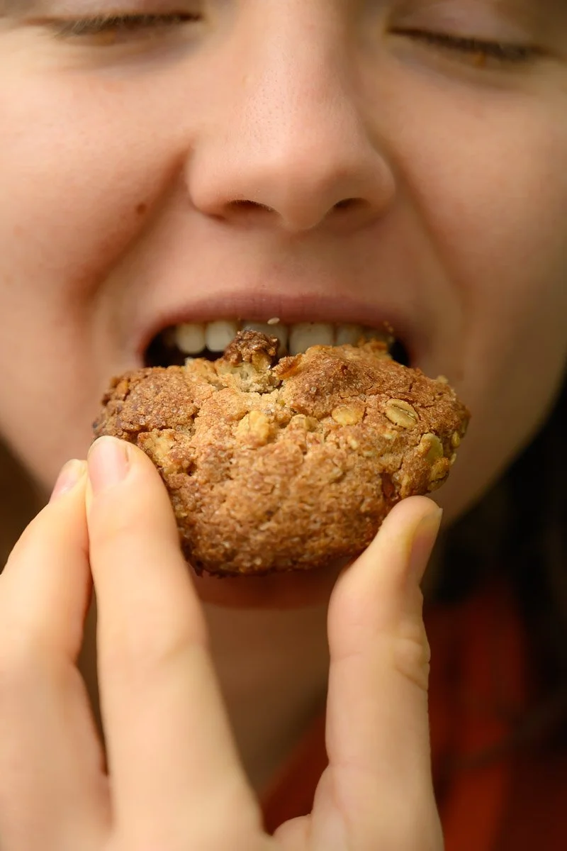 Close-up of person biting into a large chocolate chip cookie with visible oats, with closed eyes.