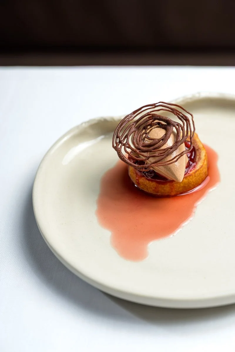 A plated dessert featuring a small cake topped with a swirl of chocolate mousse and intricate chocolate decoration, with a pink sauce drizzled on the plate.