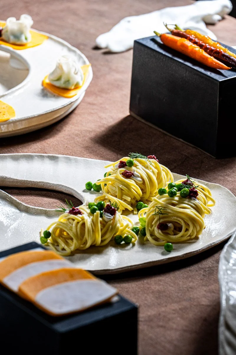 Elegant plated pasta with green peas and garnishes on a ceramic platter, surrounded by other gourmet dishes and ingredients, in a fine dining setting.