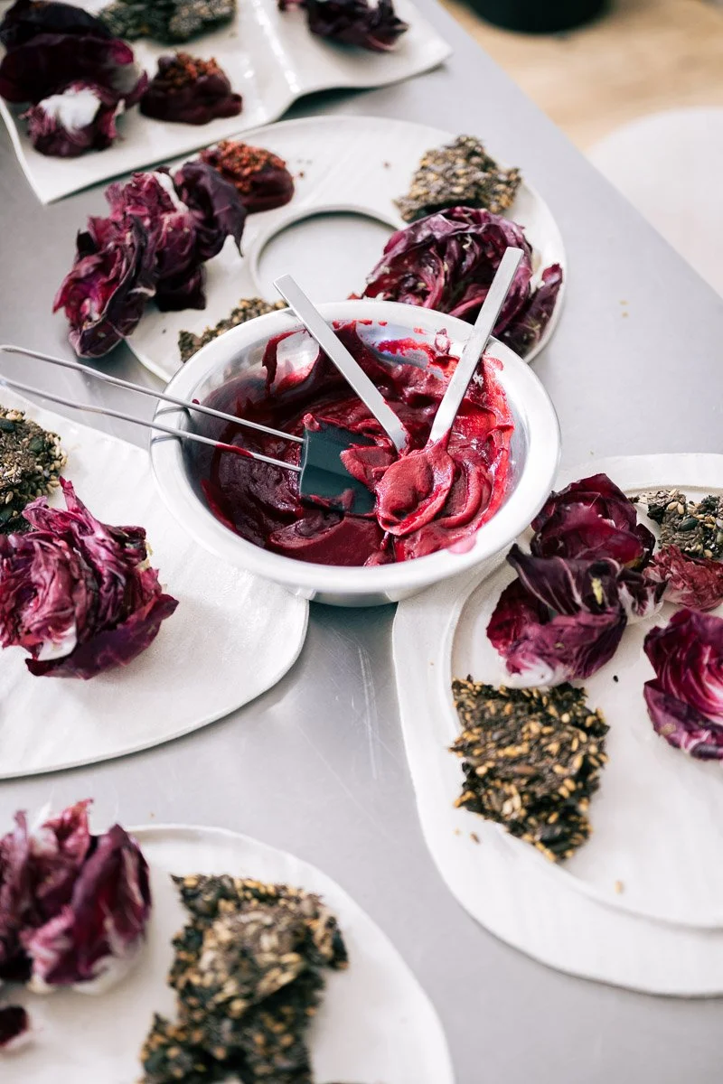 A white bowl of red berry sorbet with metal spatulas, surrounded by plates of purple leafy vegetables and sesame seed-topped crackers.