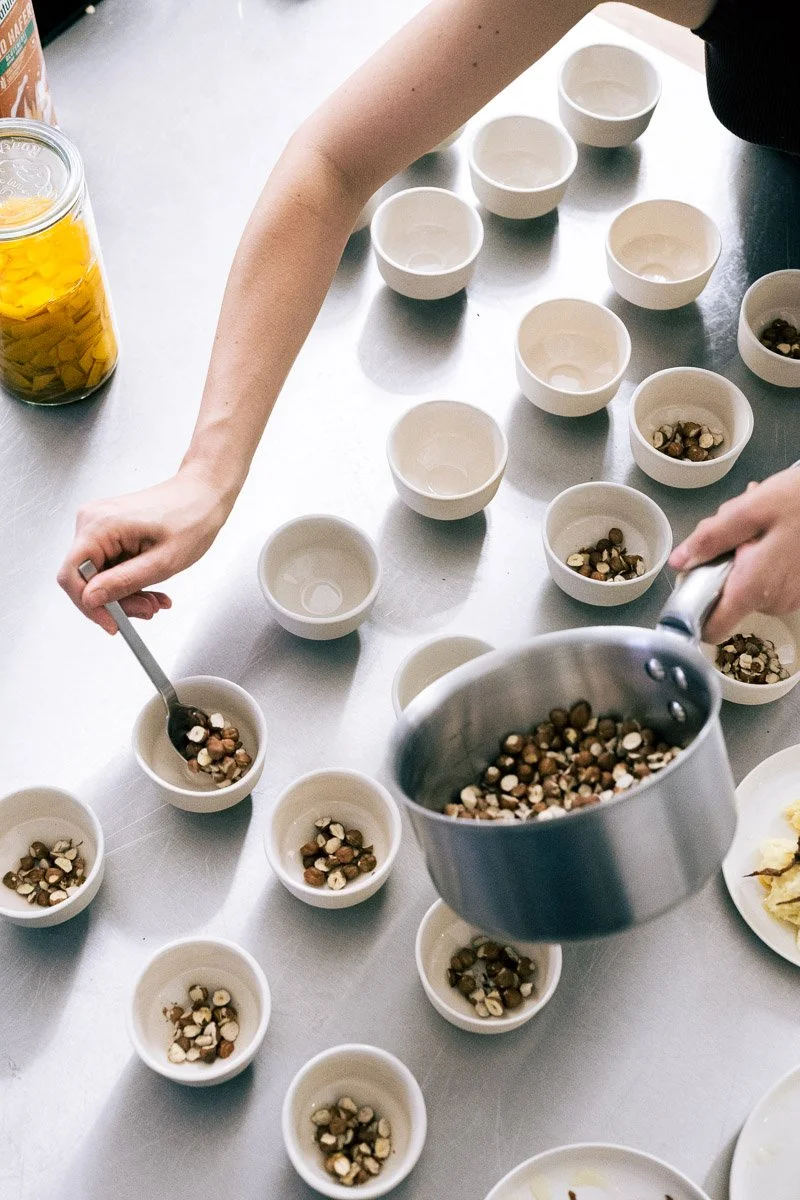 Person filling small bowls with a mixture of chopped nuts and candy, with additional bowls on a metal table.