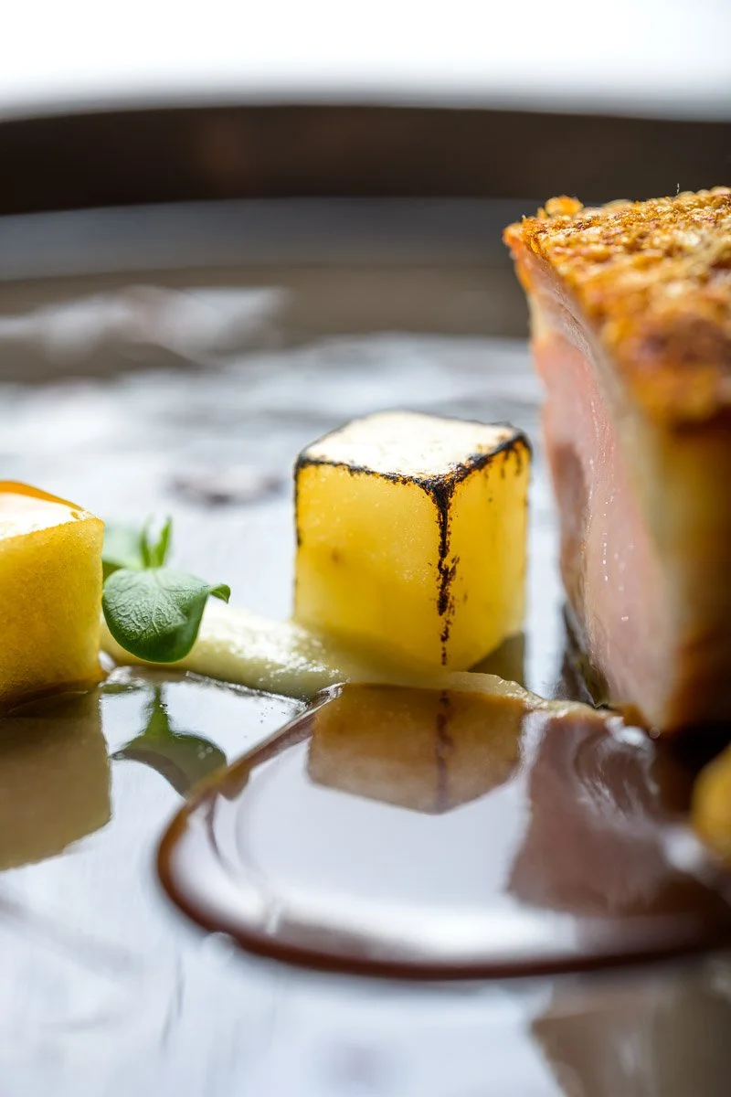 Close-up of a plated dish featuring a slice of roasted meat, a burnt cube of potato, green herbs, and a sauce.