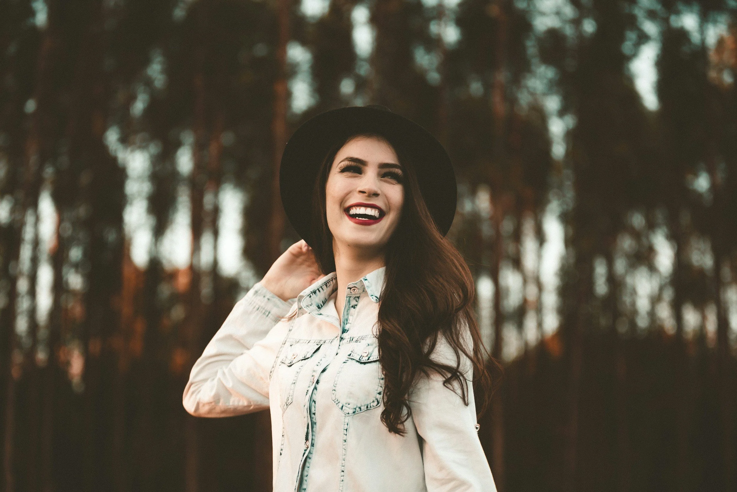 A young woman with long brown hair, smiling with red lipstick, wearing a black wide-brim hat and a casual light-colored denim button-up shirt, standing outdoors in front of a blurred forest background.