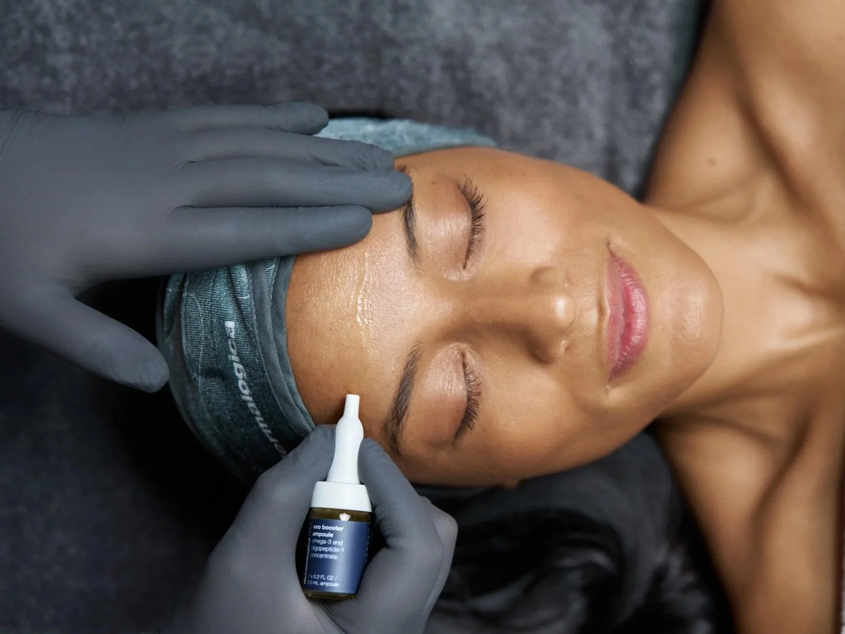 A woman receiving a facial treatment with a skincare product and a healthcare professional wearing gray gloves applying serum to her forehead.
