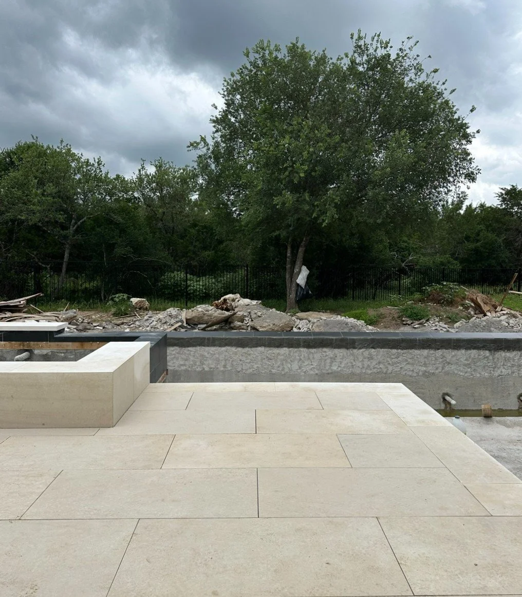 A view of an outdoor space under construction with a tiled patio in the foreground, a partially dug area with a low wall, and trees and a cloudy sky in the background.