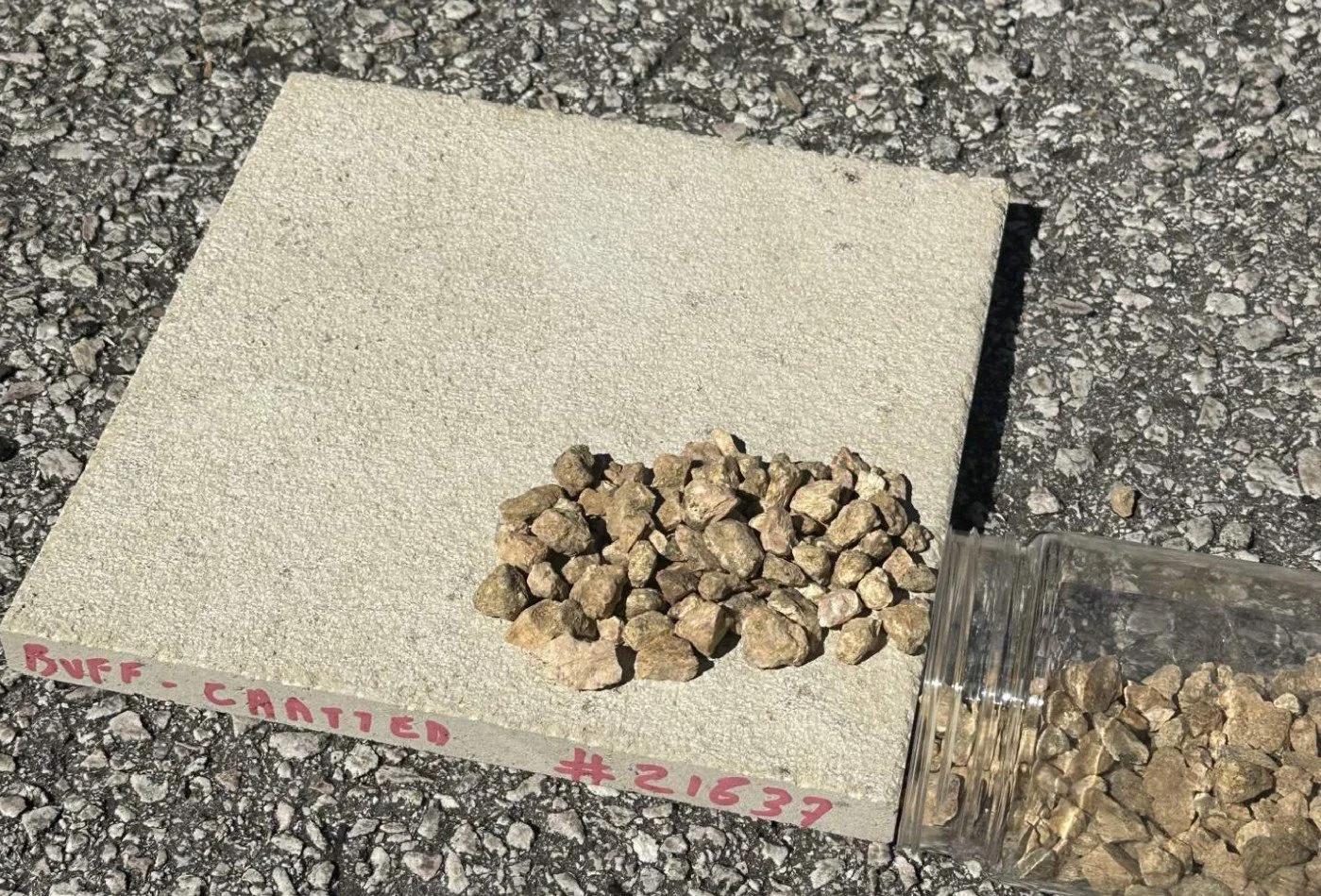 A sample of small gravel stones placed on a beige textured paper card with handwriting that says 'BUFF - CANTIED #21637', on a rough asphalt surface.