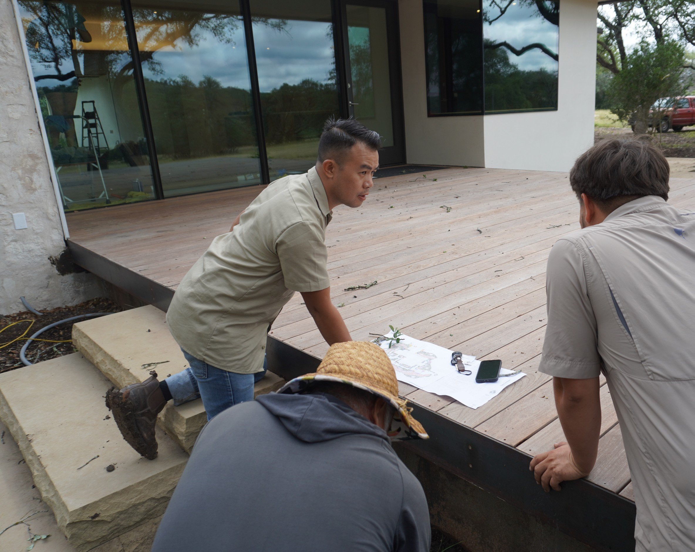 Two men are crouched down and looking at architectural plans on a table on a wooden deck, with a third person partly visible in front of them. The deck is attached to a modern building with glass doors and large windows, surrounded by trees and outdoor scenery.
