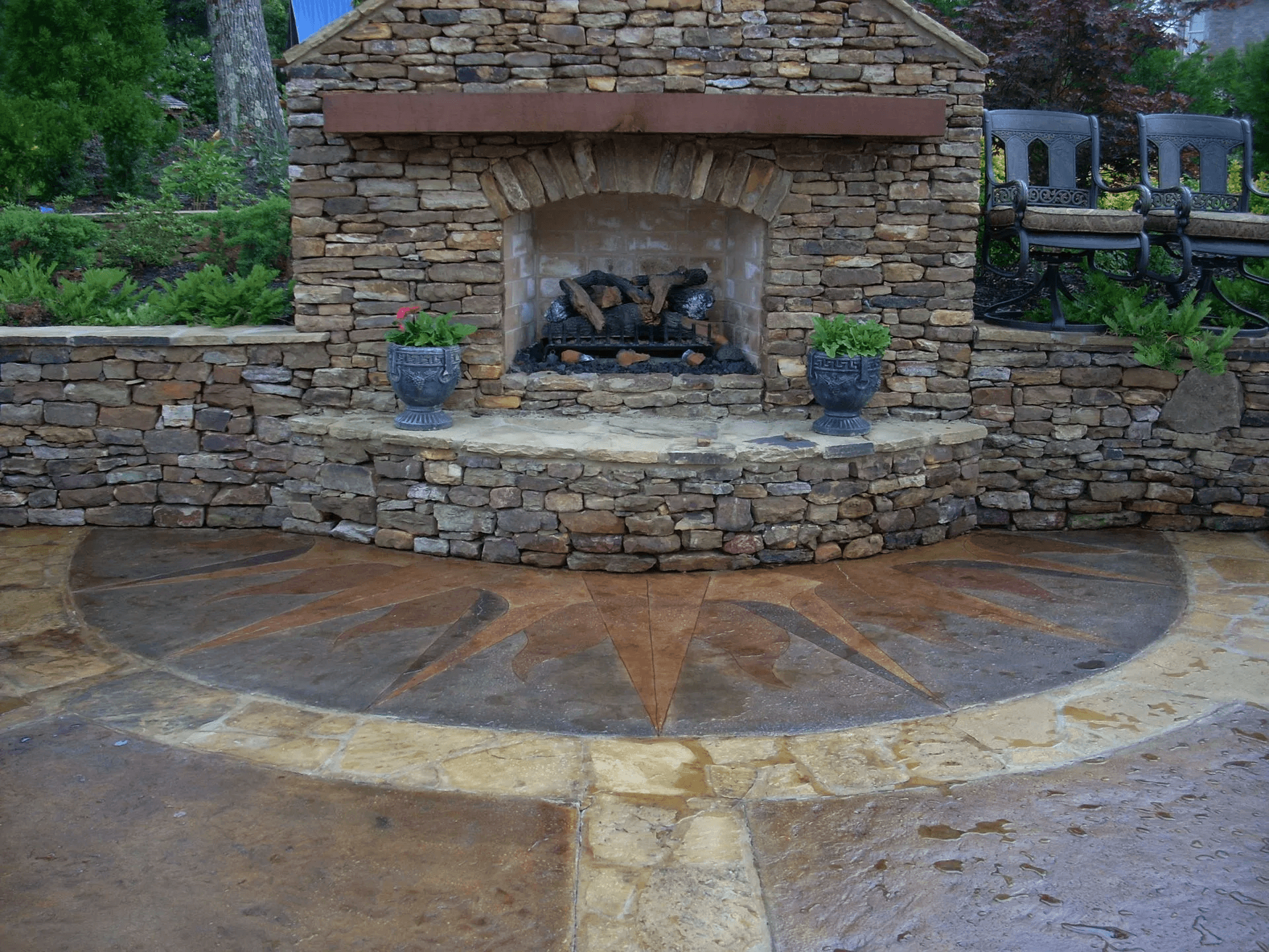 A stone outdoor fireplace with two potted plants on the edge, surrounded by a stone patio with a decorative starburst pattern.