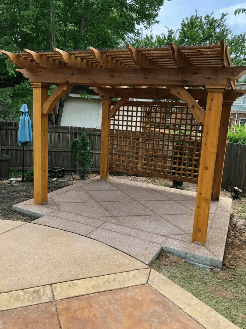 Backyard patio with a wooden pergola, lattice side panel, and concrete paving stones, surrounded by a wooden fence and trees.