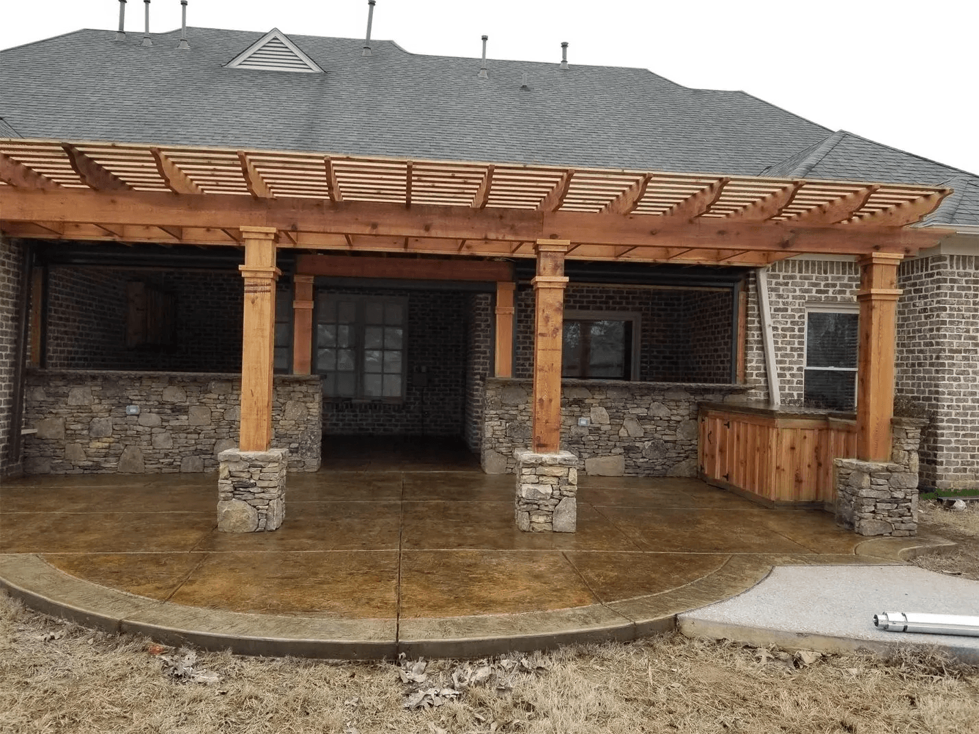 A brick house with a gray shingled roof, featuring a newly constructed wooden pergola with stone supports over a stamped concrete patio.
