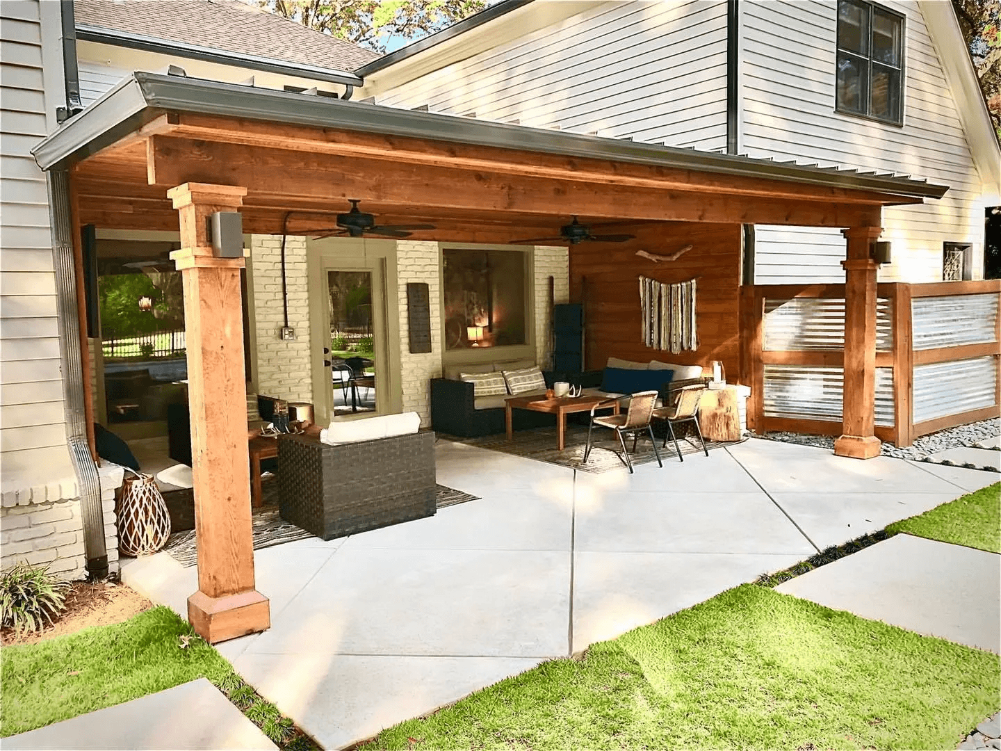 Covered outdoor patio with wooden beams, ceiling fans, and various outdoor furniture including chairs, sofas, and a coffee table, attached to a house with white siding and a brick wall.