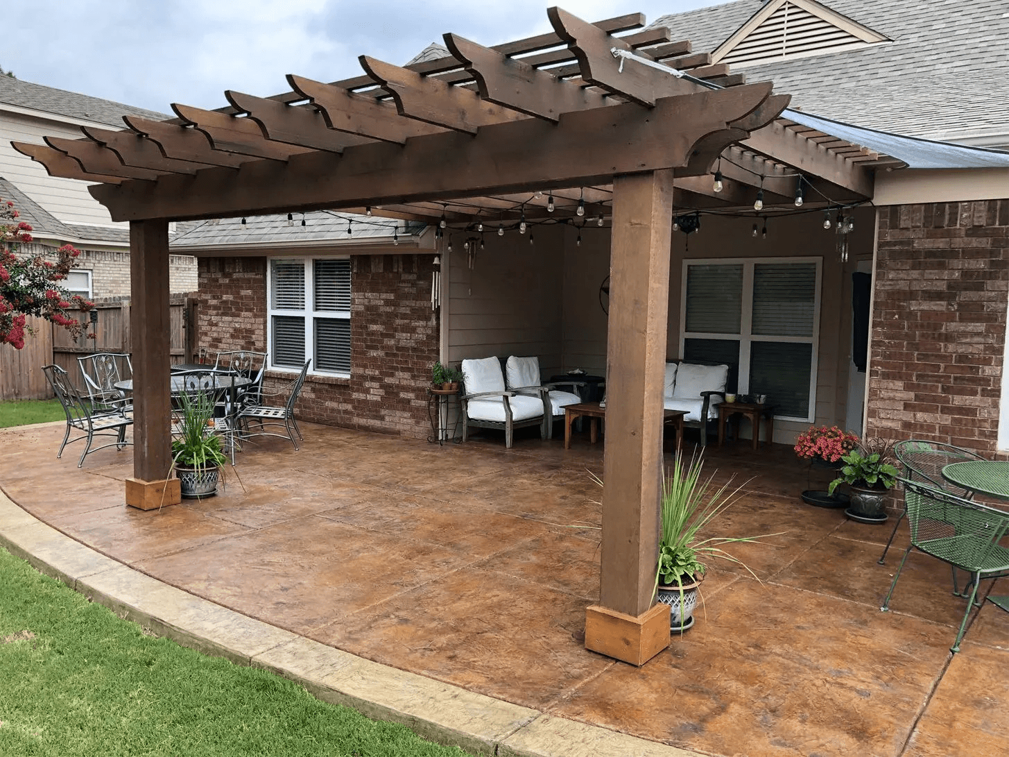 Backyard patio area with a wooden pergola overhead, outdoor furniture including a round table with chairs on the left, and white cushioned seating in the corner. Potted plants with flowers are placed around the space, with a brick house with windows in the background.