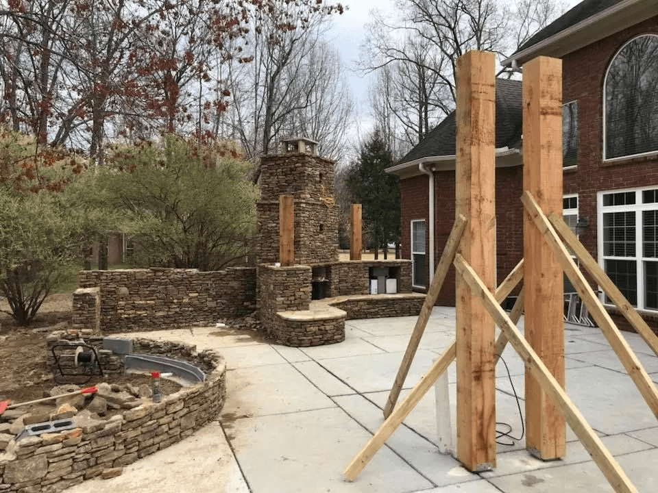 Ongoing construction of an outdoor stone fireplace and wall with wooden supports in a backyard.