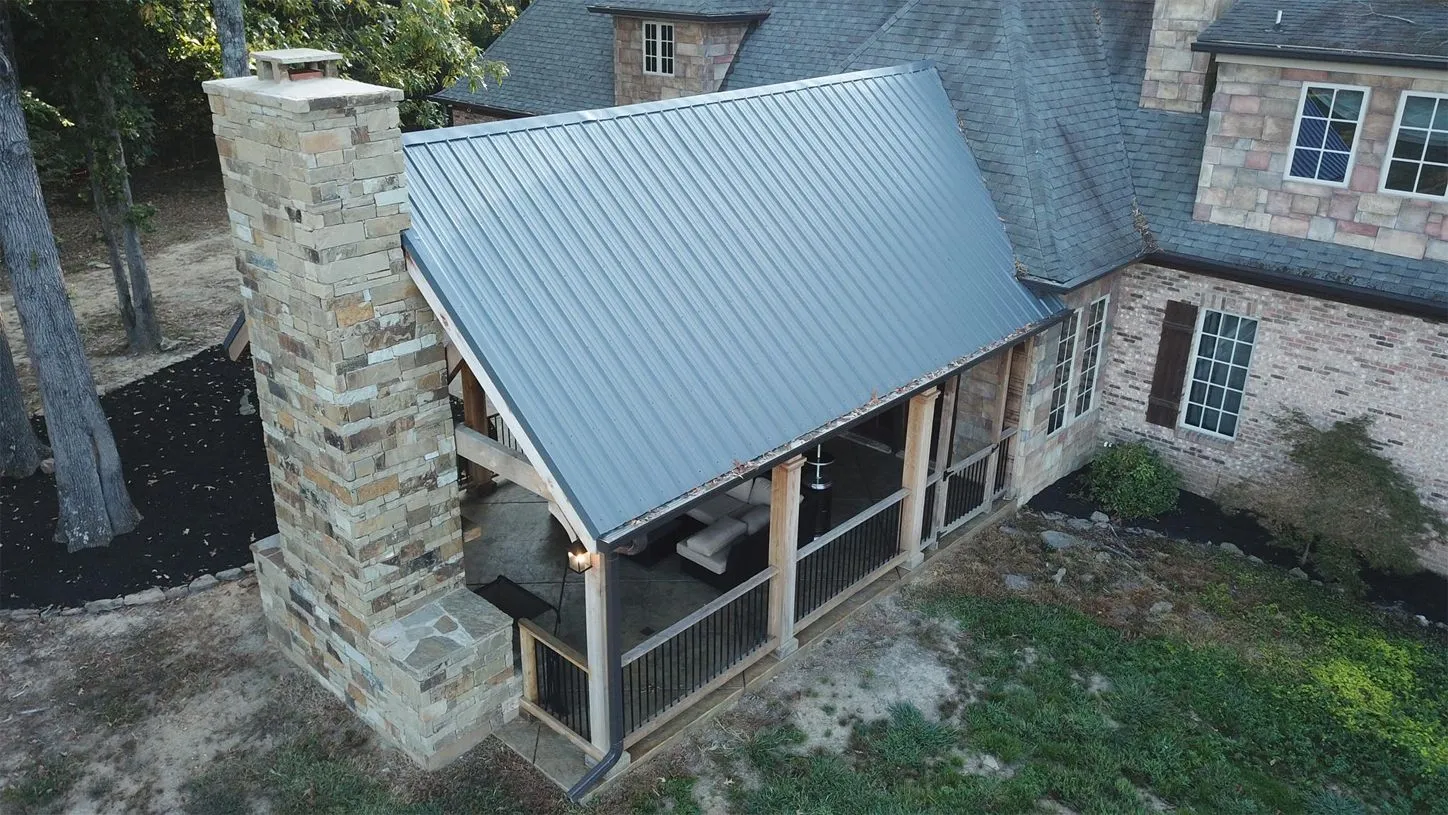 Aerial view of a house with a brick exterior, metal roofed porch, and a large stone chimney, surrounded by trees and grass.