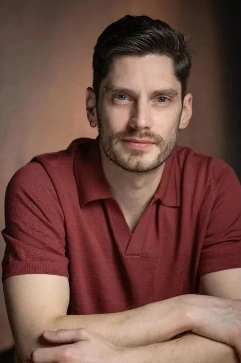 An image of Nicholas Robinson who has dark hair and a beard, wearing a red polo shirt, looking at the camera with a slight smile, seated with arms crossed on a neutral background.