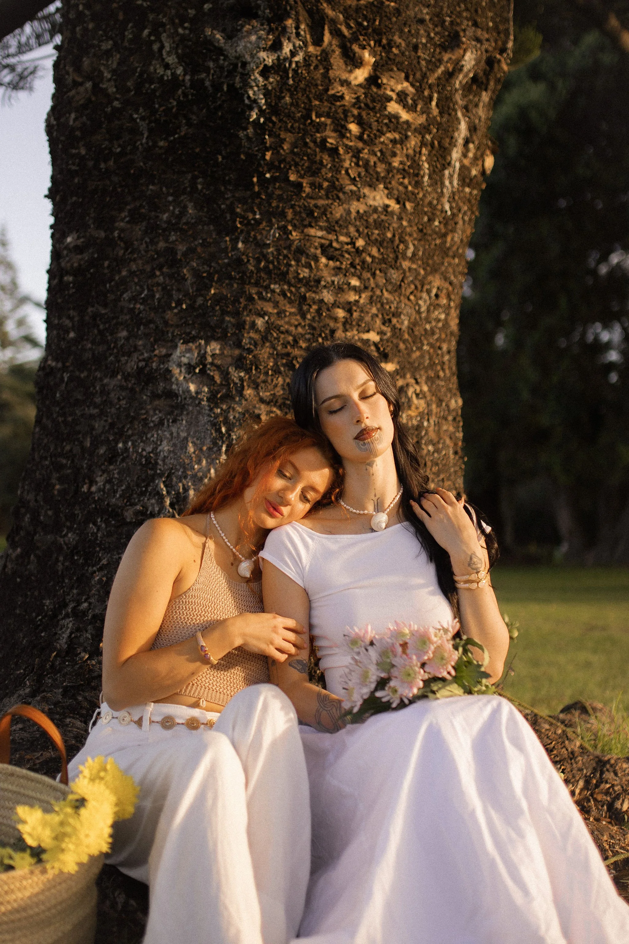 Two women sitting closely together outdoors near a large tree, one with dark hair and the other with red hair, both with closed eyes and peaceful expressions, holding flowers and leaning on each other.
