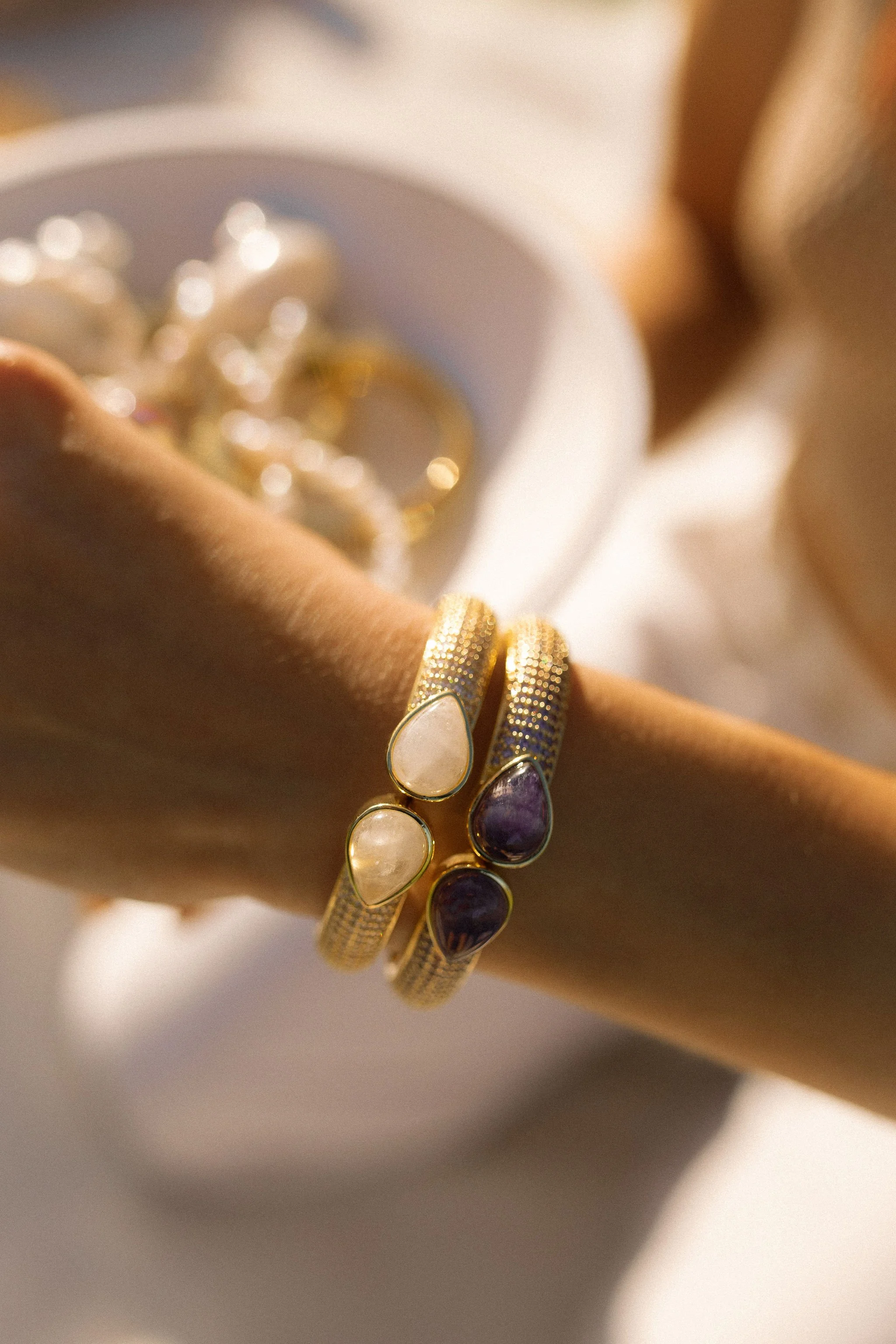 Close-up of gold bangles with white and purple stones on a person's wrist, with soft lighting.