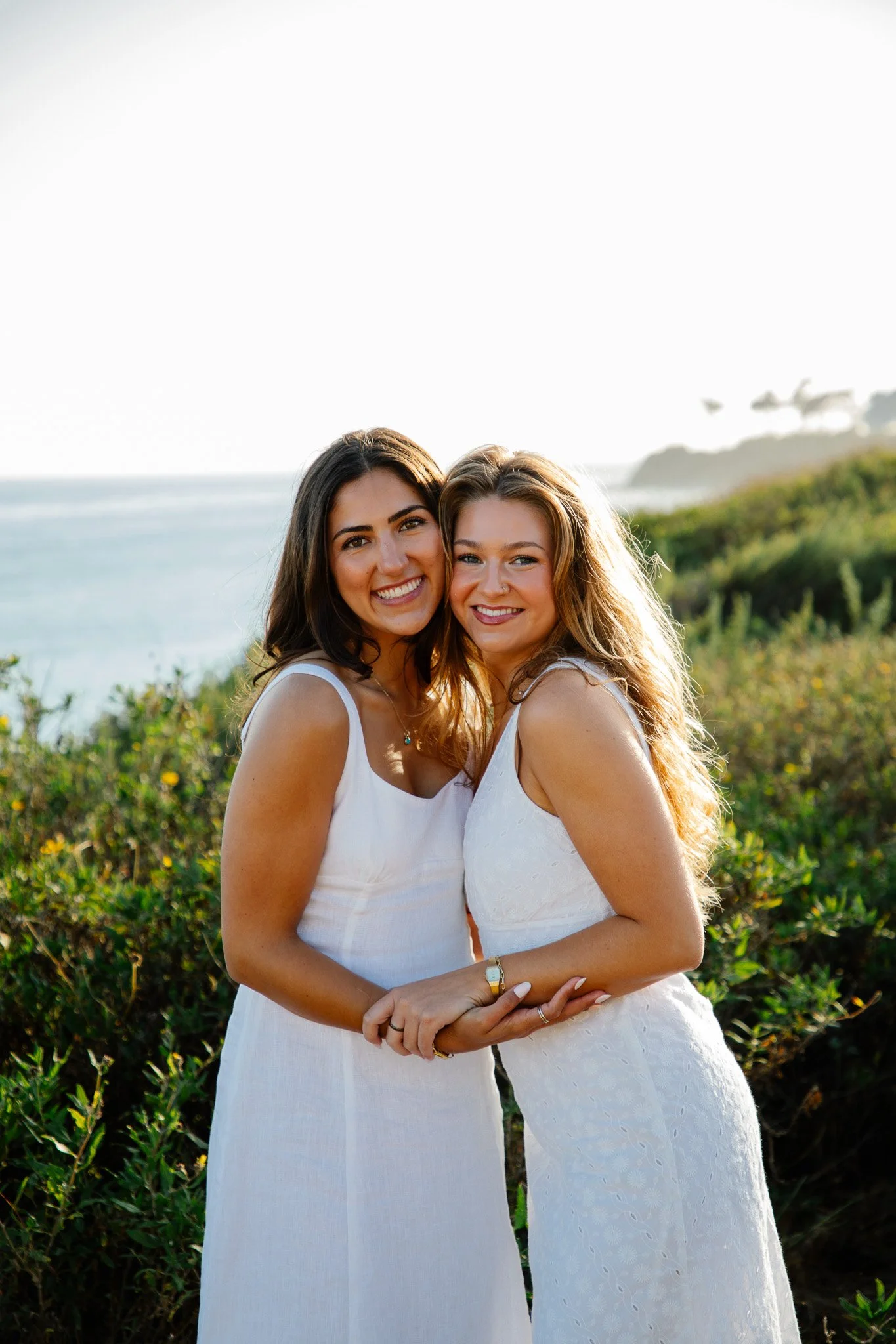 A dreamy coastal college graduation photoshoot featuring a large sorority friend group from University of California, Santa Barbara posing together in the golden evening light of Santa Barbara. The girls are dressed in coordinated white dresses and g