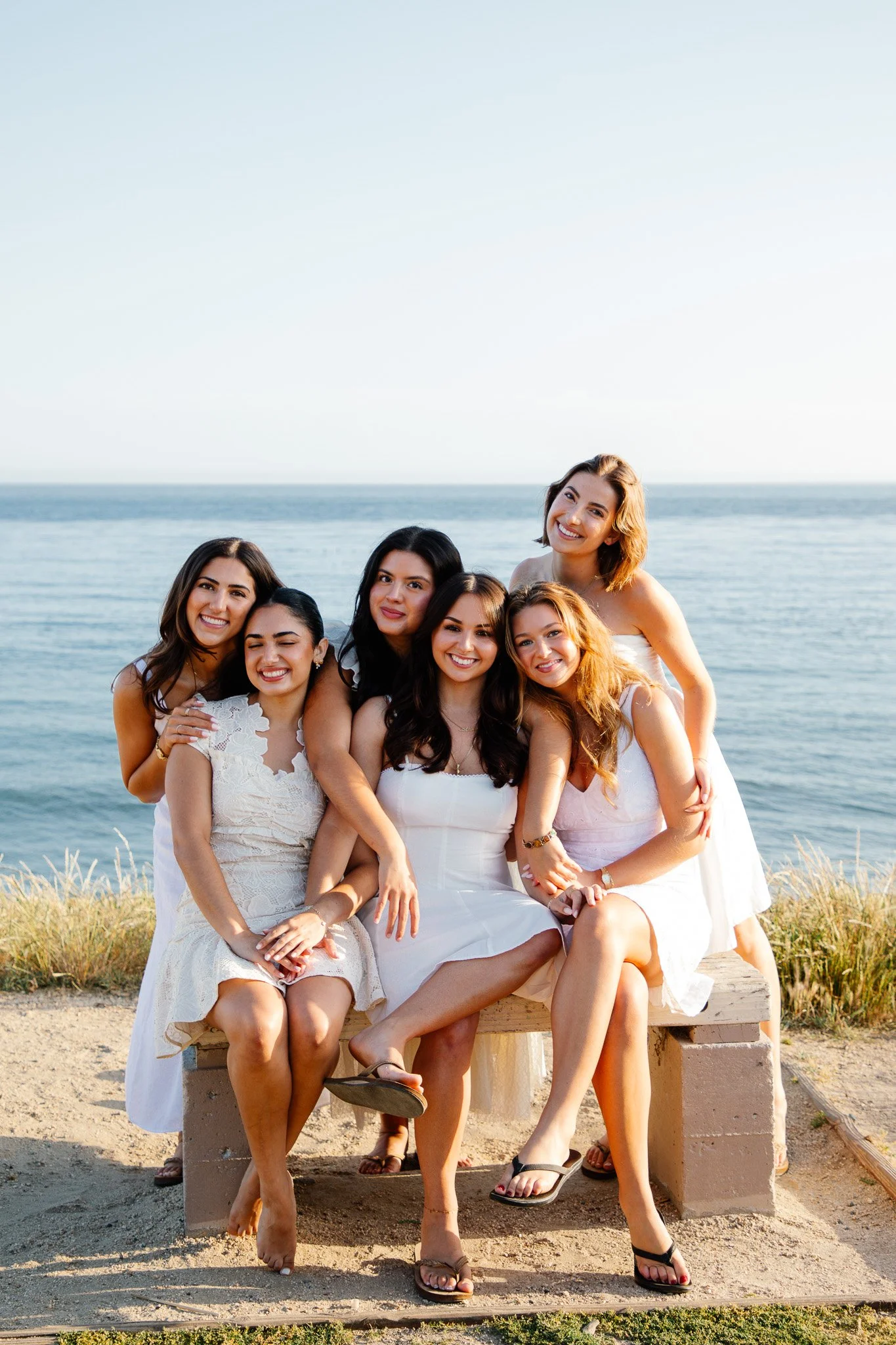 A dreamy coastal college graduation photoshoot featuring a large sorority friend group from University of California, Santa Barbara posing together in the golden evening light of Santa Barbara. The girls are dressed in coordinated white dresses and g
