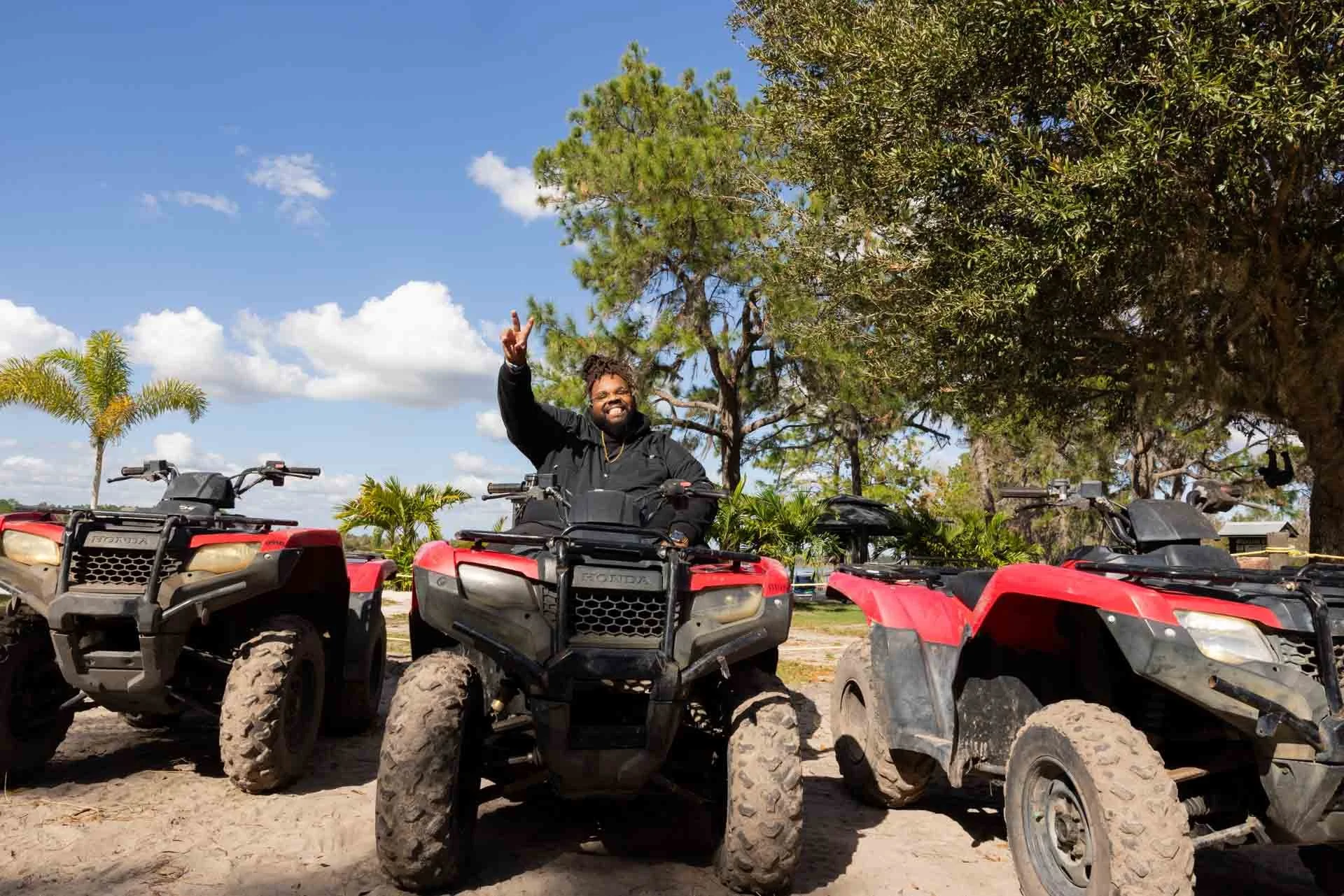 Man smiling and making a peace sign while sitting on an all-terrain vehicle (ATV) outdoors on a sunny day, with several other ATVs around him, trees, and a blue sky with clouds in the background.