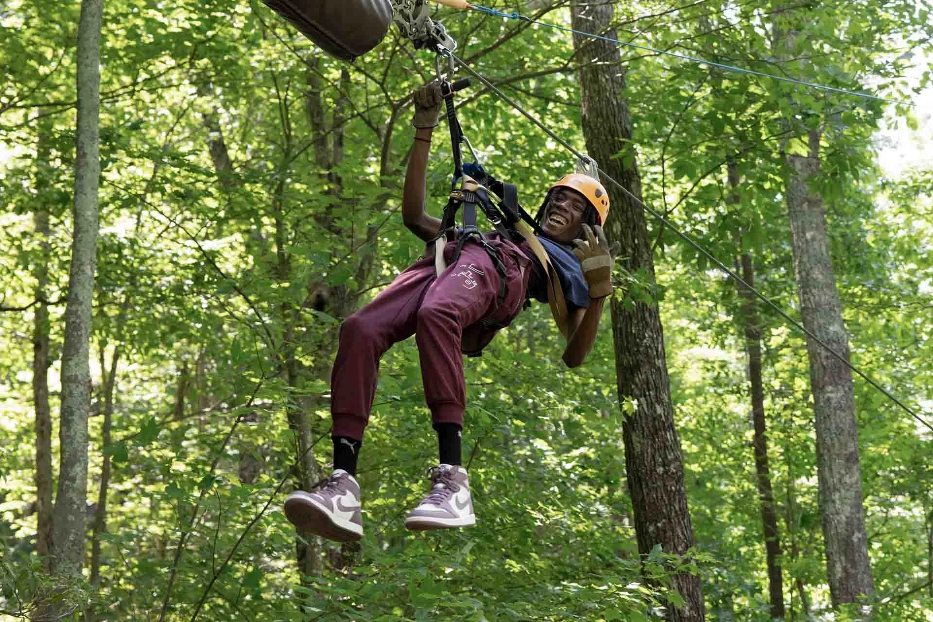 A person wearing a helmet and harness is ziplining through a green forest.