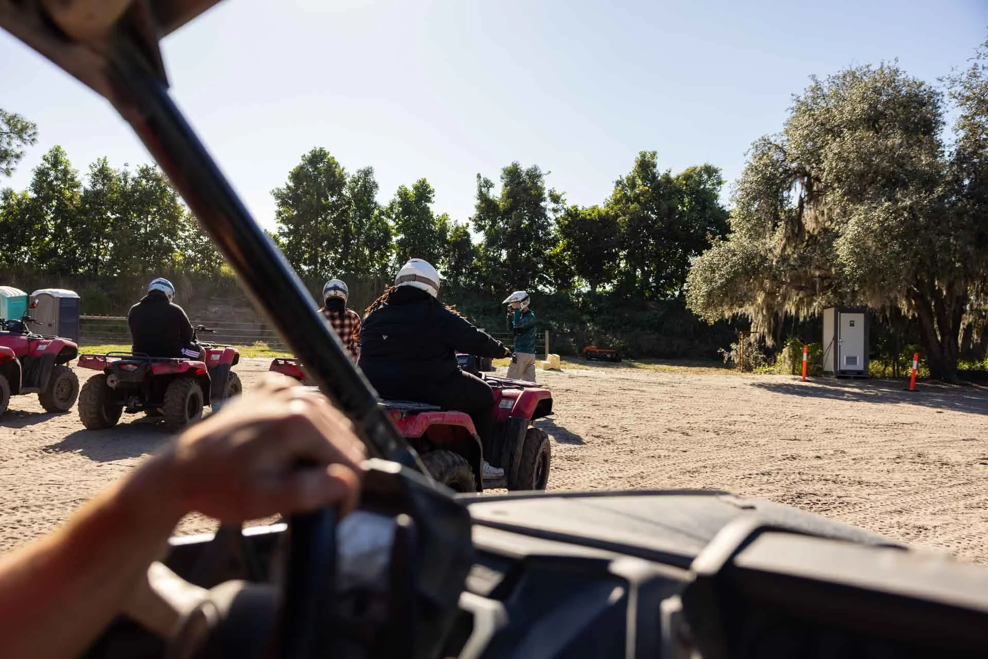 People riding ATVs in a dirt outdoor area, with some individuals wearing helmets and a person standing and talking in the background, surrounded by trees.