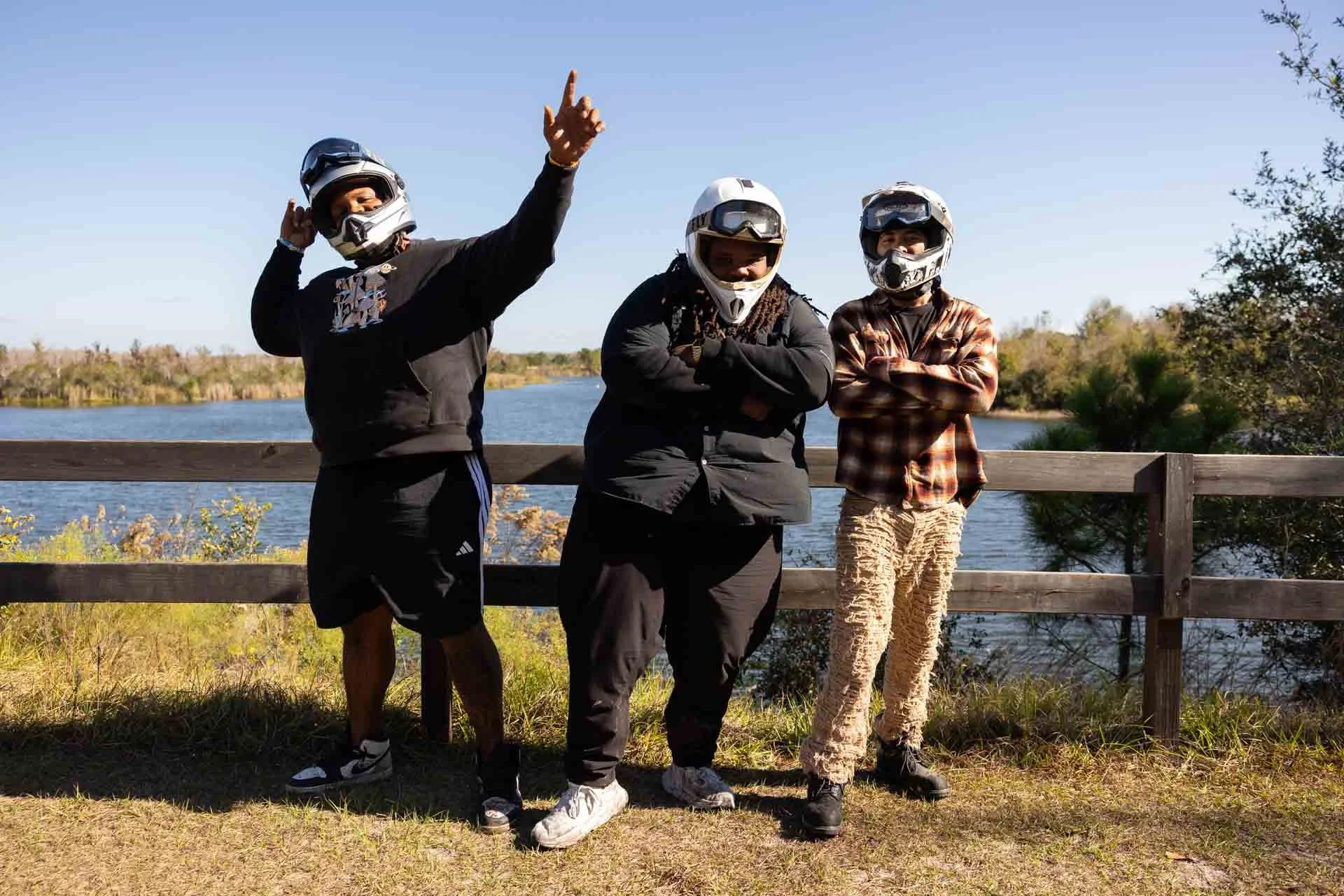 Three people wearing motorcycle helmets standing outdoors near a body of water, with a wooden railing behind them. One person is pointing upward, the others are crossing their arms.