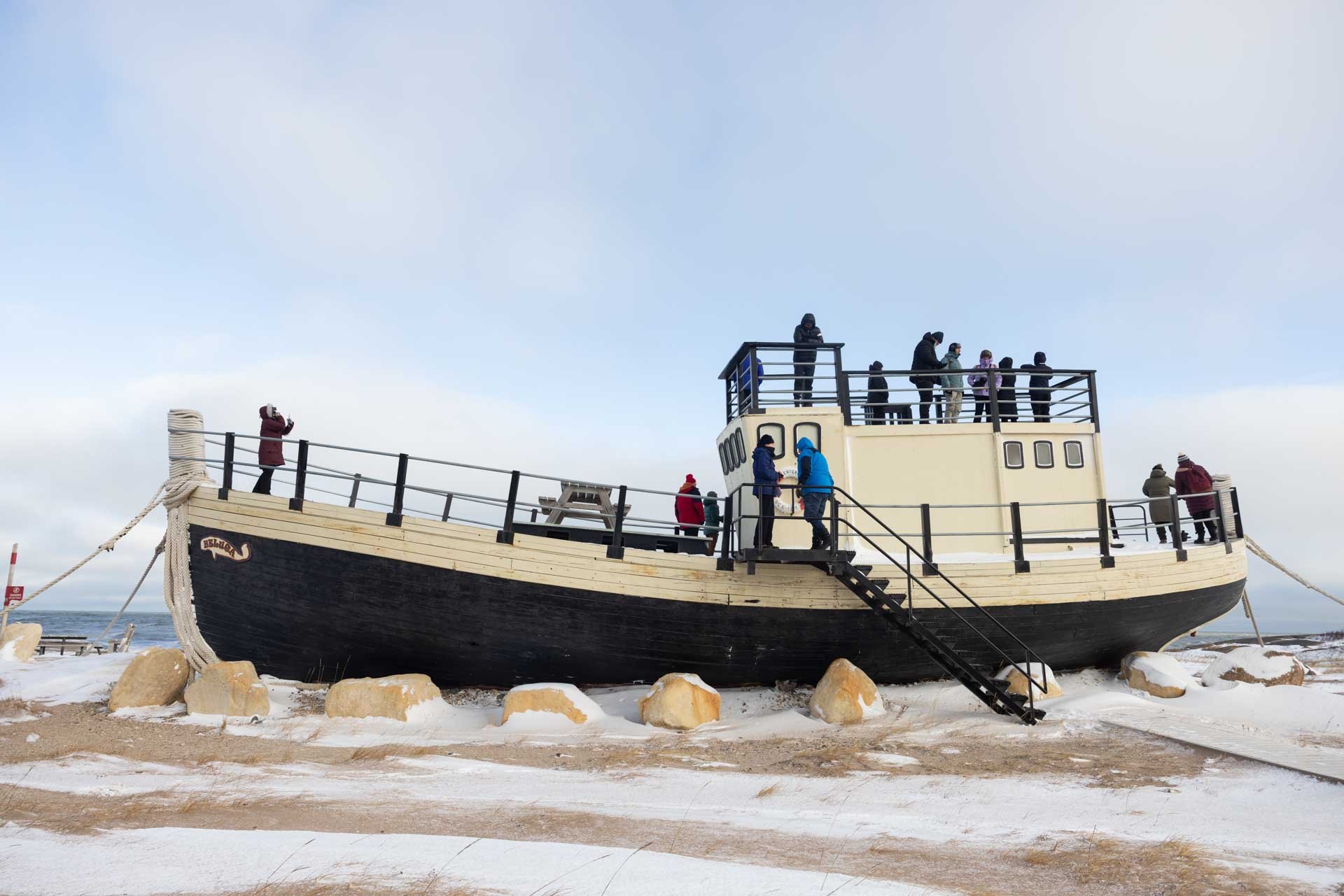A large boat-shaped structure on land with snow, rocks, and several people around it, some taking photos and others walking, under a cloudy sky.
