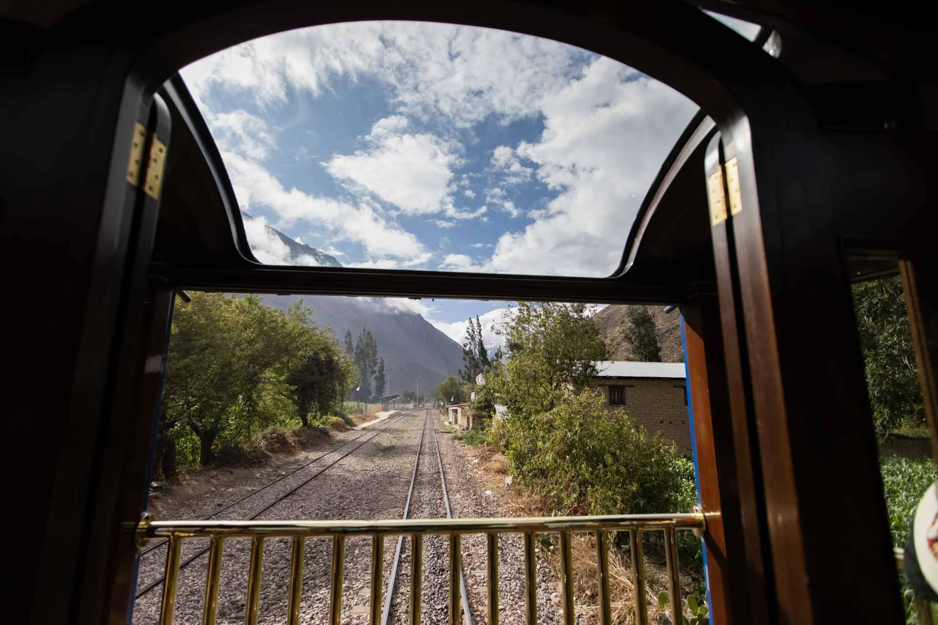 View from inside a train looking out the window at a rural landscape with trees, small buildings, train tracks, mountains, and a partly cloudy sky.