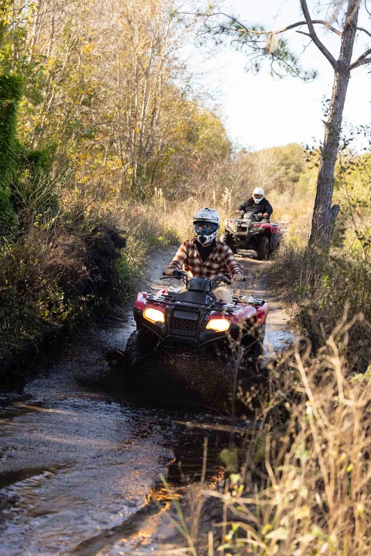 Two people riding ATVs through a muddy forest trail with trees and bushes surrounding the path.