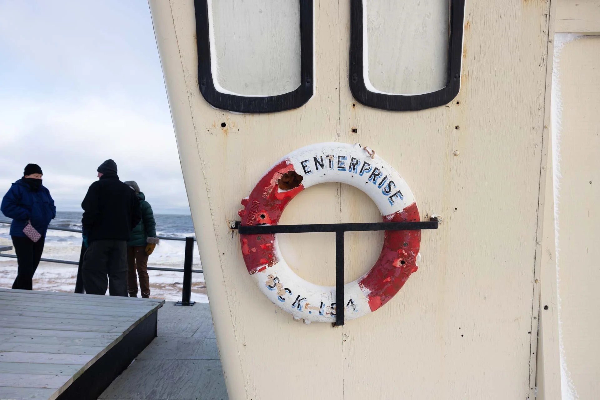 A life preserver ring with the word 'ENTERPRISE' on it is mounted on a tan wall, with a group of people standing near the ocean in the background.