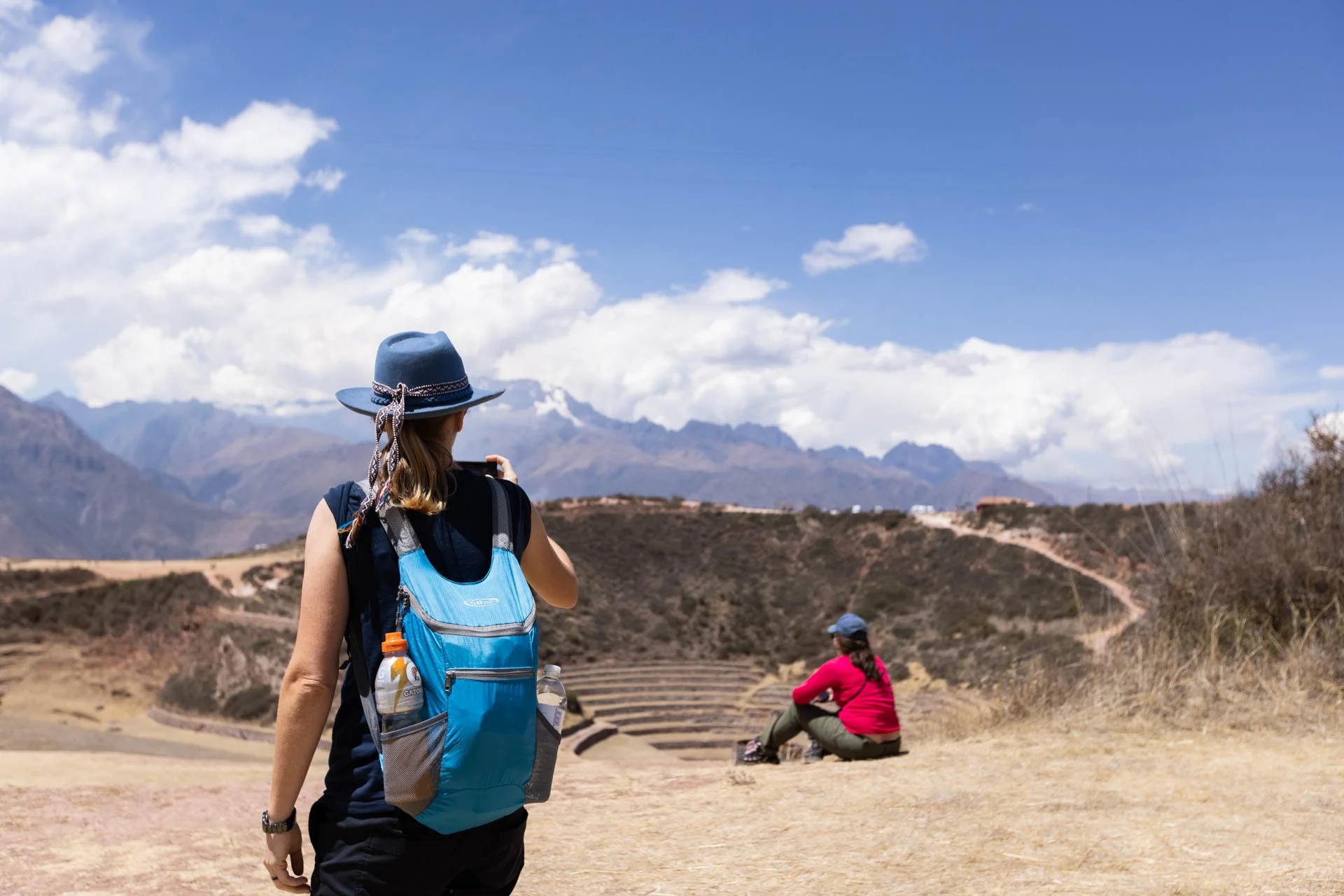 A woman with a blue hat and backpack takes a photo while another woman sitting on the ground looks at the mountains in a dry, hilly landscape under a partly cloudy sky.